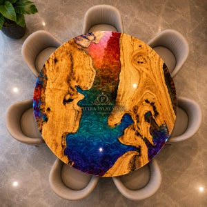 May include: A round dining table featuring a natural wood slab with a central river design made of swirling blue, teal, and red resin. The table is surrounded by six curved, light grey upholstered chairs. The logo PIETRA INLAY STONE is visible.