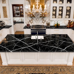 May include: A kitchen island featuring a black marble countertop with white veining and a built-in white sink with a silver faucet. The island base is white with recessed paneling, set against a backdrop of white cabinetry and a crystal chandelier.