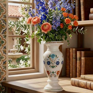 May include: A white marble vase with a flared rim and base, featuring intricate floral inlay patterns in blue, orange, and green. The vase is filled with a vibrant bouquet of blue delphiniums, orange ranunculus, and green eucalyptus leaves.