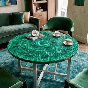 Puede incluir: Una mesa de comedor redonda con un tablero de patrón de malaquita verde vibrante y acabado pulido, sostenida por una base de metal plateado con un marco en forma de X. La superficie muestra intrincados patrones de bandas concéntricas.