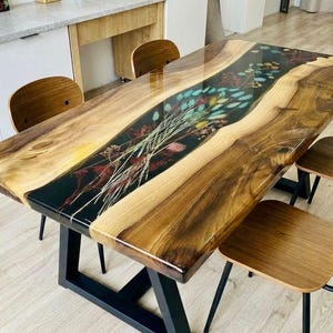 Può includere: Un tavolo da pranzo rettangolare con due lastre di legno naturale a bordo vivo unite da un inserto centrale in resina nera. La sezione in resina contiene fiori e piante essiccate. Il tavolo ha una struttura in metallo nero e quattro sedie in legno.