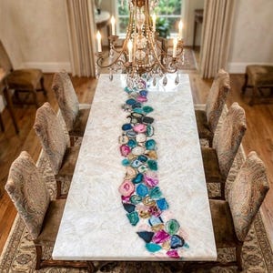 May include: A rectangular dining table with a white surface and a colorful agate stone centerpiece. The table is surrounded by eight upholstered chairs and a decorative chandelier hangs above. The room has hardwood floors and a patterned rug.