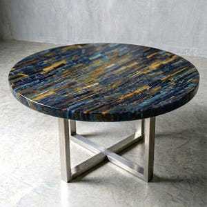 May include: A round coffee table featuring a polished blue and gold tiger eye gemstone surface with a linear, multi-directional pattern. The tabletop is supported by a silver-colored metal base constructed with four intersecting legs forming an X-shape.
