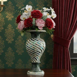 Puede incluir: Un jarrón decorativo con un patrón de rayas contiene un ramo de rosas rojas, peonías rosas y orquídeas blancas. El jarrón está sobre una mesa de madera oscura, contra un papel pintado verde estampado y una cortina burdeos.