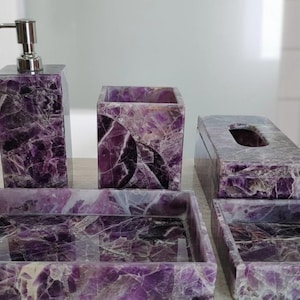 Può includere: Un set da bagno con un dosatore di sapone con pompa argentata, un portaspazzolino, un portatovaglioli e due vassoi. Il set è realizzato in pietra marmorizzata viola e bianca.
