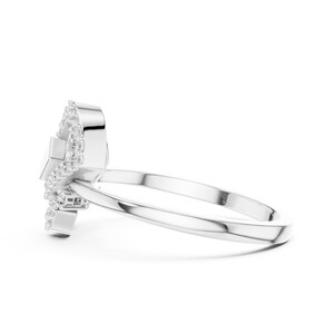 May include: A silver ring with a unique design, featuring a central square-shaped stone surrounded by a halo of smaller, sparkling stones. The ring's band is a simple, polished silver. The ring is set against a white background.