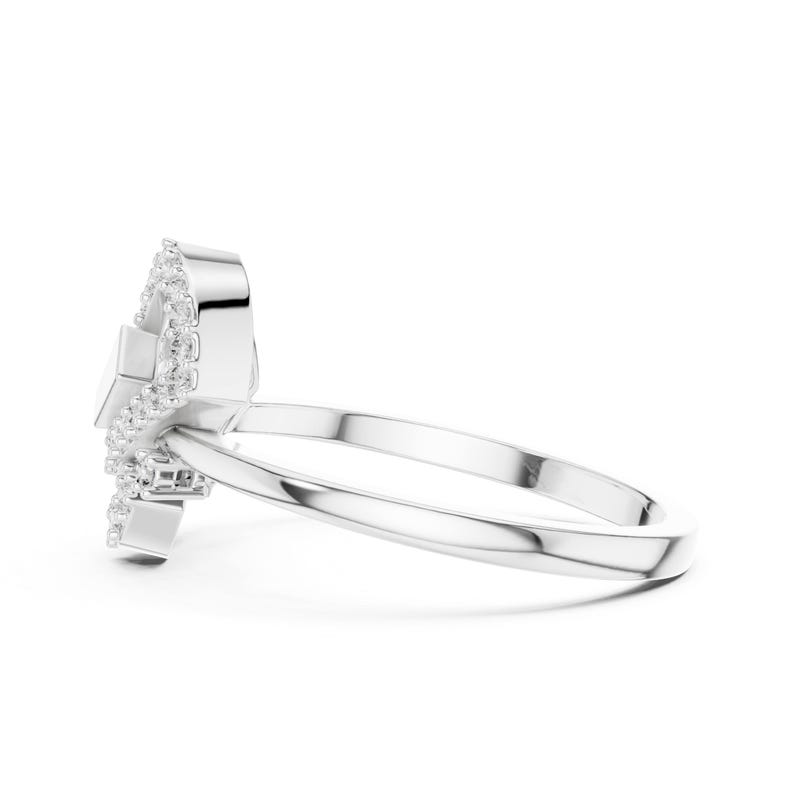 May include: A silver ring with a unique design, featuring a central square-shaped stone surrounded by a halo of smaller, sparkling stones. The ring's band is a simple, polished silver. The ring is set against a white background.
