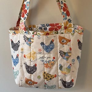 May include: A quilted tote bag with a cream background featuring colorful chickens. The bag has floral patterned straps and trim. The text "just for you" is on a small patch on the front of the bag. The bag is hanging on a wooden wall hook.