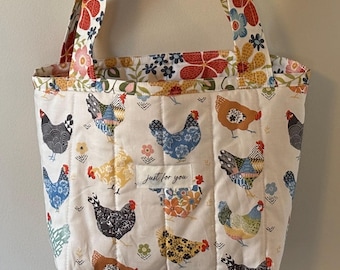 Könnte beinhalten: Eine gesteppte Tragetasche mit cremefarbenem Hintergrund und bunten Hühnern. Die Tasche hat Träger und Verzierungen mit Blumenmuster. Der Text "just for you" befindet sich auf einem kleinen Aufnäher auf der Vorderseite der Tasche. Die Tasche hängt an einem hölzernen Wandhaken.