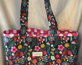 Könnte beinhalten: Eine Tragetasche mit einem dunkelgrünen Blumenmuster mit Pilzen, Blumen und Schmetterlingen. Die Tasche hat ein rosa gepunktetes Futter und Griffe mit dem gleichen floralen Design. Ein kleines Etikett lautet "just for you."