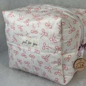 May include: A white quilted cosmetic bag with a repeating pattern of pink bows. The bag has a zipper closure and a small tag that says "just for you". A wooden tag with "handmade by Gobin" hangs from the zipper.