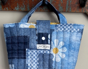 May include: A blue denim tote bag with a patchwork design. The bag features daisies, checkered patterns, and solid blue denim. It has a top handle, a flap closure with a white button, and the text "just for you" on a small label.