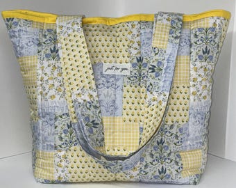 Könnte beinhalten: Eine gesteppte Tragetasche mit einem Patchwork-Design aus gelben und blauen Blumen- und Vichy-Karomustern. Die Tasche hat einen leuchtend gelben Rand an der Oberkante und zwei passende Schulterriemen. Ein weißes Stoffetikett trägt die Aufschrift just for you.