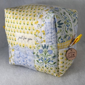May include: A quilted fabric cosmetic bag with a patchwork design. The bag features yellow floral and blue floral patterns, with a zipper closure and a small tag that says "just for you". A wooden tag with the name "Goblin" is attached.