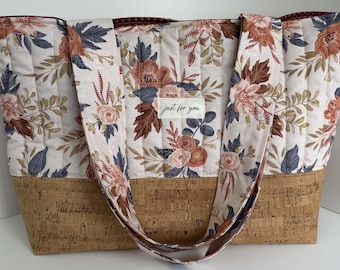 Könnte beinhalten: Eine gesteppte Tragetasche mit Blumenmuster in Altrosa, Braun und Marineblau auf cremefarbenem Grund. Der Boden besteht aus natürlichem Korkmaterial. Ein rechteckiges weißes Etikett auf der Vorderseite zeigt den Text Just for you.
