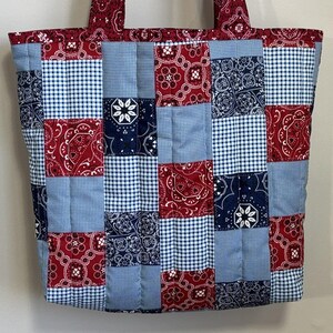 May include: A patchwork tote bag with a quilted design. The bag features a mix of red paisley, blue gingham, and navy blue floral patterns. The handles and top border are red paisley. The bag is a medium size.