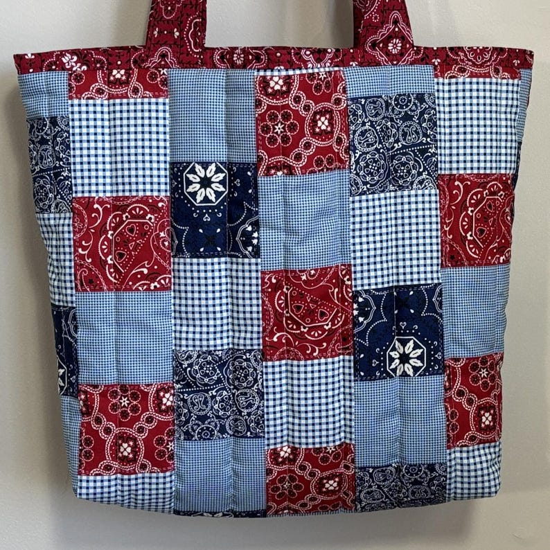 May include: A patchwork tote bag with a quilted design. The bag features a mix of red paisley, blue gingham, and navy blue floral patterns. The handles and top border are red paisley. The bag is a medium size.