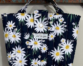 Könnte beinhalten: Eine gesteppte Tragetasche mit marineblauem Hintergrund und einem weißen Gänseblümchen-Blumenmuster. Die Tasche hat zwei Griffe und einen Knopfverschluss. Der Text "just for you" steht auf einem kleinen weißen Etikett.
