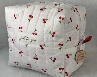 May include: A white, quilted cosmetic bag featuring a cherry print and the text "just for you." It has a zip closure and a wooden tag reading "handmade by Gobin."