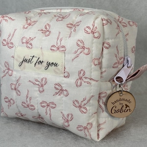May include: A quilted, off-white cosmetic bag with a repeating pattern of pink bows. The bag features a zipper closure and a small tag that says "just for you." A wooden charm with the text "handmade by Gobin" hangs from the zipper.