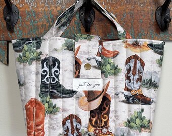 Könnte beinhalten: Gesteppte Tragetasche mit Western-Motiv. Die Tasche hat ein sich wiederholendes Muster aus Cowboy-Stiefeln, Hüten und Kakteen auf hellem Hintergrund. Die Tasche hat einen Griff und ein kleines Etikett mit der Aufschrift "just for you".