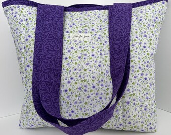 Könnte beinhalten: Eine große Tragetasche mit einem Blumenmuster aus kleinen lila Blumen und grünen Blättern auf weißem Grund. Die Tasche hat breite, einfarbige lila Träger und Besätze. Der Text "just for you" ist auf der Vorderseite aufgedruckt.