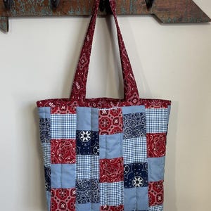 May include: A patchwork tote bag with a quilted design. The bag combines red paisley, blue gingham, and navy blue floral patterns. The handles and top trim are red paisley. The bag is suspended from a wooden arrow-shaped wall hook.
