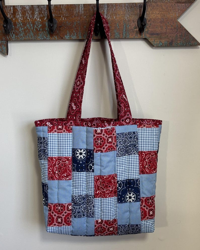 May include: A patchwork tote bag with a quilted design. The bag combines red paisley, blue gingham, and navy blue floral patterns. The handles and top trim are red paisley. The bag is suspended from a wooden arrow-shaped wall hook.