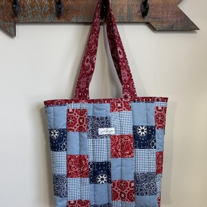 May include: A patchwork tote bag in red, white, and blue. The bag has a variety of patterns, including paisley and gingham. The handles and top border are a solid red paisley. The bag is hanging on a wooden arrow-shaped coat rack. The bag is made of quilted fabric.