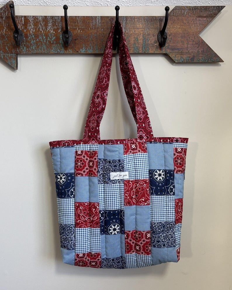 May include: A patchwork tote bag in red, white, and blue. The bag has a variety of patterns, including paisley and gingham. The handles and top border are a solid red paisley. The bag is hanging on a wooden arrow-shaped coat rack. The bag is made of quilted fabric.