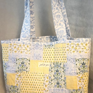 May include: A patchwork tote bag with a floral and gingham pattern in yellow and blue. The bag features long handles and a label that reads "just for you". The bag is made of quilted fabric.