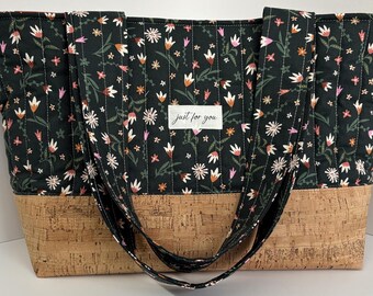 Könnte beinhalten: Eine gesteppte Tragetasche mit einem schwarzen Stoffoberteil mit mehrfarbigem Blumenmuster und einem naturfarbenen Korkboden. Die Tasche verfügt über zwei passende Stoffschulterriemen und ein weißes rechteckiges Etikett mit dem Text just for you.