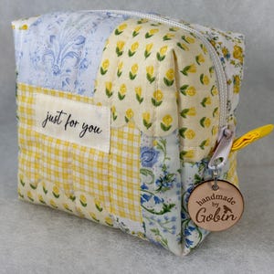 May include: A patchwork cosmetic bag with a white zipper and a yellow pull tab. The bag features floral and gingham patterns in yellow and blue. A tag reads "just for you" and a wooden tag says "handmade by Gobin".