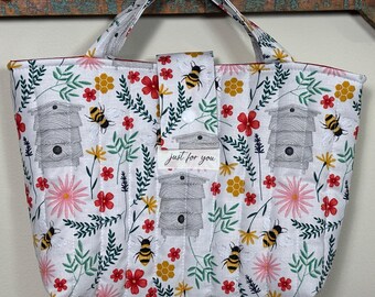 Könnte beinhalten: Eine weiße, gesteppte Tragetasche mit einem Blumen- und Bienenmuster. Das Muster umfasst rote, gelbe und rosa Blumen, Bienenstöcke und Bienen. Die Tasche hat einen Griff und einen Druckknopfverschluss mit dem Text "just for you".