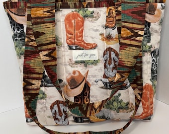 Könnte beinhalten: Gesteppte Tragetasche mit Western-Motiv. Die Tasche zeigt Bilder von Cowboy-Stiefeln und -Hüten auf cremefarbenem Hintergrund. Die Griffe und der Besatz haben ein Muster in Braun-, Grün- und Orangetönen. Der Text "just for you" befindet sich auf der Vorderseite.