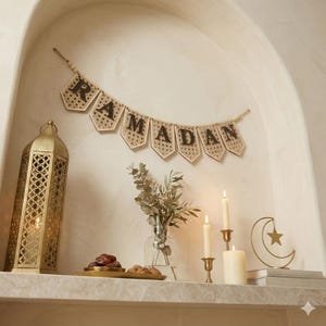 Pu&ograve; includere: Un banner decorativo del Ramadan con dettagli intrecciati &egrave; appeso sopra una mensola. Sono esposti una lanterna dorata, candele e una luna crescente con una stella. Un vaso di verde e un vassoio di datteri completano l'allestimento festivo.