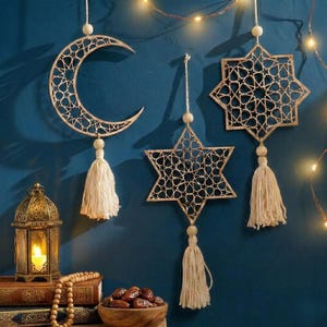 May include: Decorative Ramadan ornaments in the shape of a crescent moon, a star, and an eight-pointed star, all with intricate openwork designs. Each ornament hangs from a string and features a tassel. A lit lantern and dates in a wooden bowl are also visible.