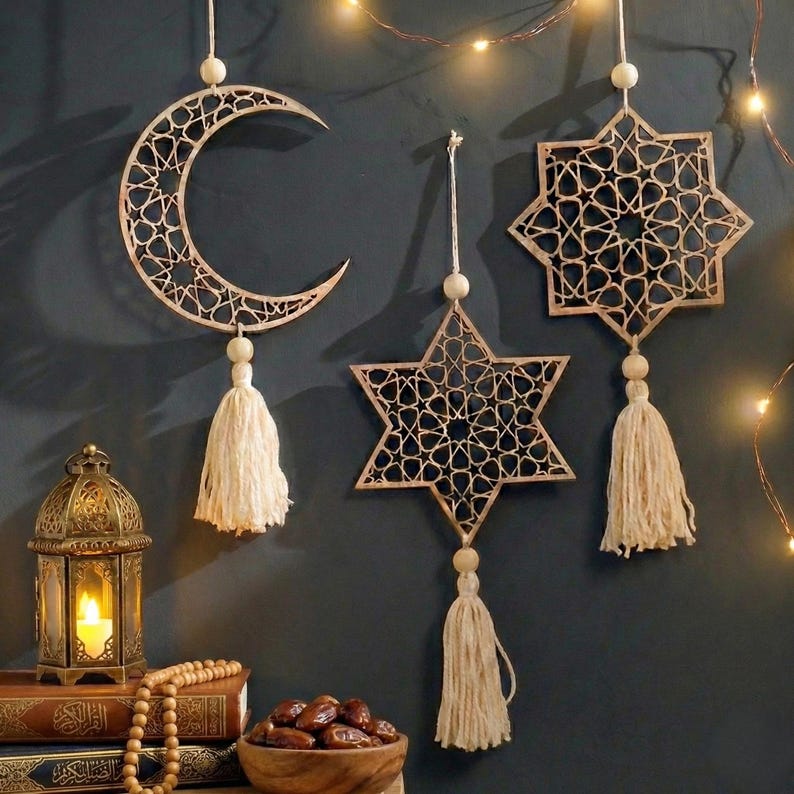 May include: Wooden decorative hanging ornaments in the shape of a crescent moon, a six-pointed star, and an eight-pointed star. Each ornament has a detailed cutout design and a tassel. The ornaments are beige and hang against a dark blue wall.