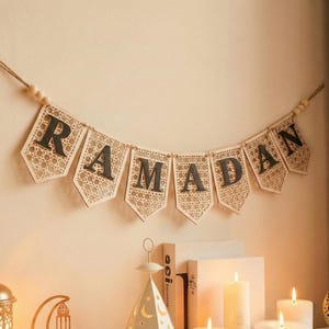 Pu&ograve; includere: Un banner del Ramadan con la parola "RAMADAN" scritta su singole bandierine. Le bandierine sono beige con un motivo intrecciato e lettere grigio scuro. Il banner &egrave; appeso a uno spago con perline di legno, adatto per le feste.