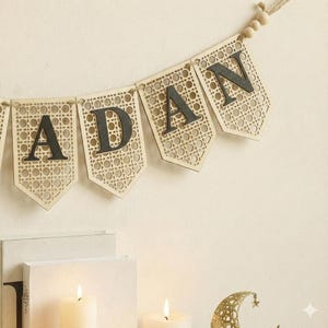 Pu&ograve; includere: Un banner di legno con la scritta "ADAN" con lettere grigio scuro. Lo striscione &egrave; realizzato in legno chiaro con un motivo intrecciato. Lo striscione &egrave; infilato con spago naturale e perline di legno. Due candele accese e una luna crescente decorativa sono in primo piano.