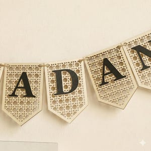 Pu&ograve; includere: Uno striscione di legno che riporta la parola "ADAM" in lettere nere. Ogni lettera &egrave; su un gagliardetto separato con un motivo intrecciato simile alla canna. Lo striscione &egrave; infilato su uno spago naturale.