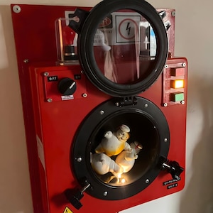 Può includere: Una scatola di metallo rossa con una finestra circolare, contenente tre figure di ceramica. La scatola ha una cornice circolare nera, una piccola finestra con una lampadina e vari interruttori e simboli di avvertimento. Il testo "CONTAINMENT UNIT 3" è visibile.