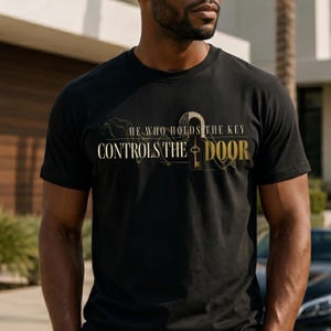 Könnte beinhalten: Ein schwarzes Kurzarm-T-Shirt mit einem gold-weißen Grafikdesign, das den Text "HE WHO HOLDS THE KEY CONTROLS THE DOOR" und eine in den Buchstaben O integrierte Schlüsselillustration zeigt, vor einem Muster aus feinen, geschwungenen Linien.