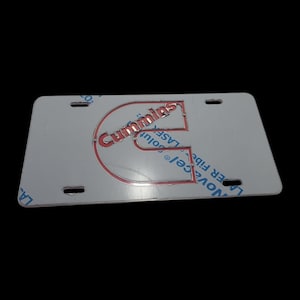 May include: A rectangular, silver-colored license plate with the red "Cummins" logo and text. The plate has four pre-drilled holes for mounting.