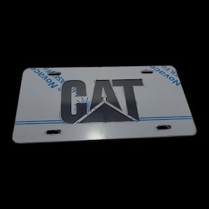 May include: A silver license plate with the CAT logo in dark gray. The plate has four mounting holes and is rectangular. The CAT logo is a stylized representation of the company's name and logo.