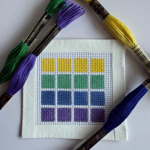 May include: A square needlepoint canvas with a grid pattern in yellow, green, blue, and purple. Several skeins of embroidery floss in matching colors surround the canvas, including green, purple, yellow, and blue.