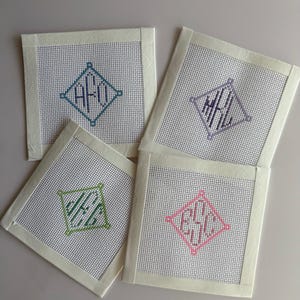 May include: Four square needlepoint coasters with personalised monogram designs. Each coaster features a diamond-shaped monogram in different colours: teal, purple, green, and pink. The coasters have a cream-coloured border.