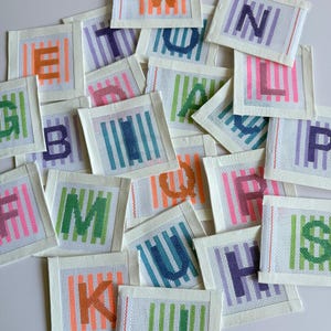 Puede incluir: Una colección de parches de letras de punto de aguja en varios colores. Cada parche presenta una letra diferente, cosida en tonos vibrantes como naranja, verde y morado, con rayas verticales. Los parches están sobre un fondo blanco.