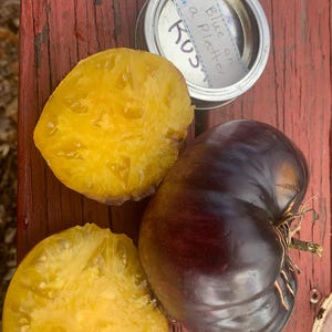 May include: A whole dark purple tomato next to two bright yellow tomato slices. A small round metal tin is present with a white label reading "Blue on a Platter" and "Kus". The items are arranged together on a red wooden surface.
