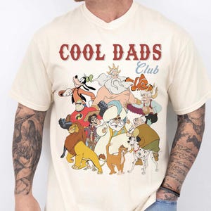 May include: Cream-colored t-shirt with the text "COOL DADS Club" in red and white. The shirt features a colorful graphic of cartoon characters. The shirt is made of a soft material.